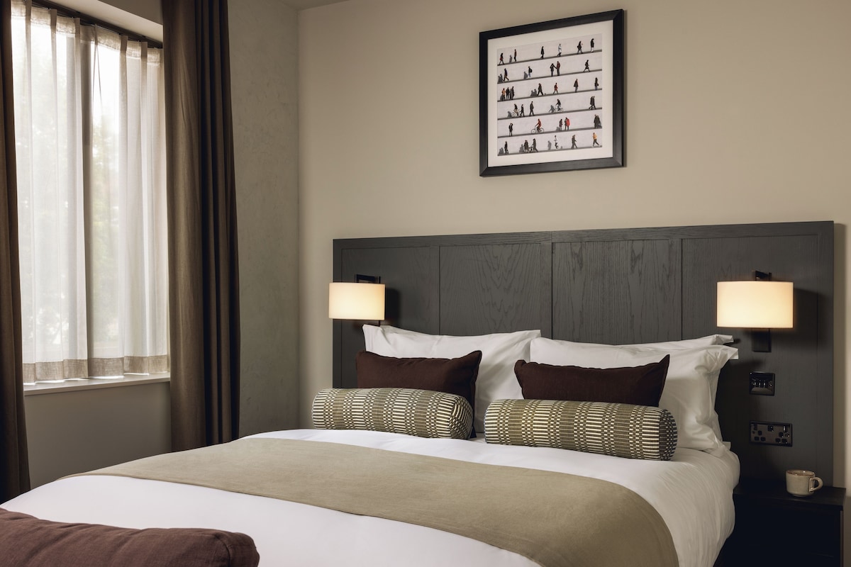 A hotel room with a bed, two bedside lamps, a framed artwork on the wall, and a window with curtains.
