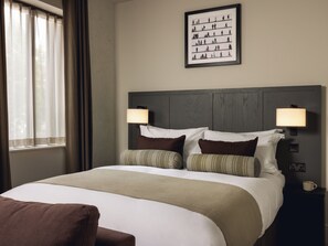 Una camera d'albergo con un letto, due lampade ai comodini, un quadro appeso al muro e una finestra con tende.