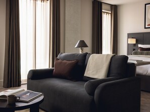 Una moderna camera d'albergo con un divano scuro, un comodino con una lampada e un letto con testiera.