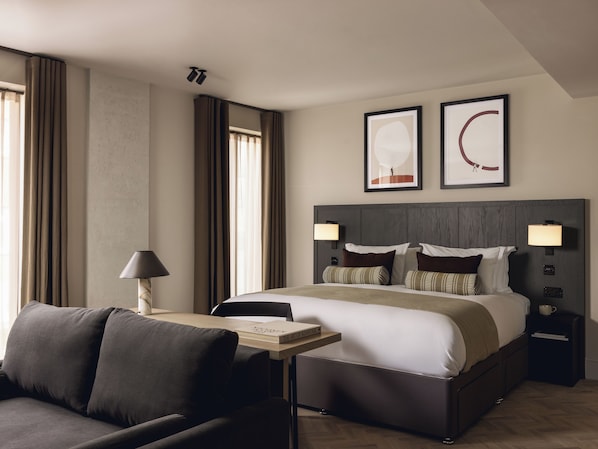 Habitación de hotel moderna con una cama grande, una mesita de noche con lámpara, una silla y vistas a la ciudad.
