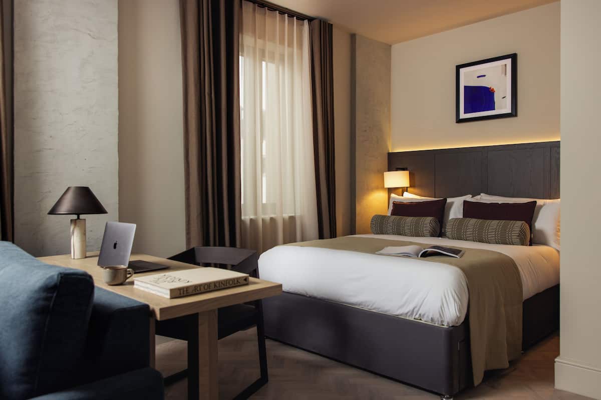 A hotel room with a bed, a desk with a laptop, a chair, a lamp, and a framed picture on the wall.