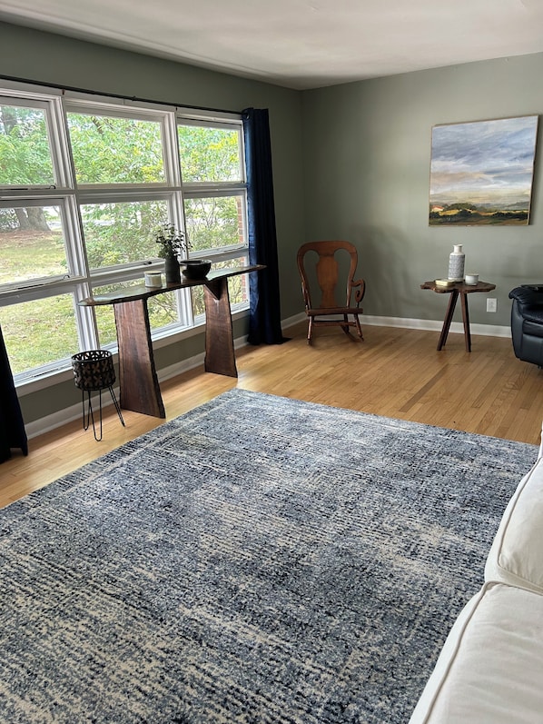 Ein Zimmer mit großem Fenster, Holzboden, blauem Teppich, einem Sessel, einem kleinen Tisch mit einer Vase und einem Gemälde an der Wand.