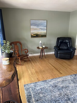 Ein Wohnzimmer mit einem Holzschaukelstuhl, einem schwarzen Ledersessel, einem kleinen Tisch mit Vase und Kerze sowie einem Gemälde an der Wand.