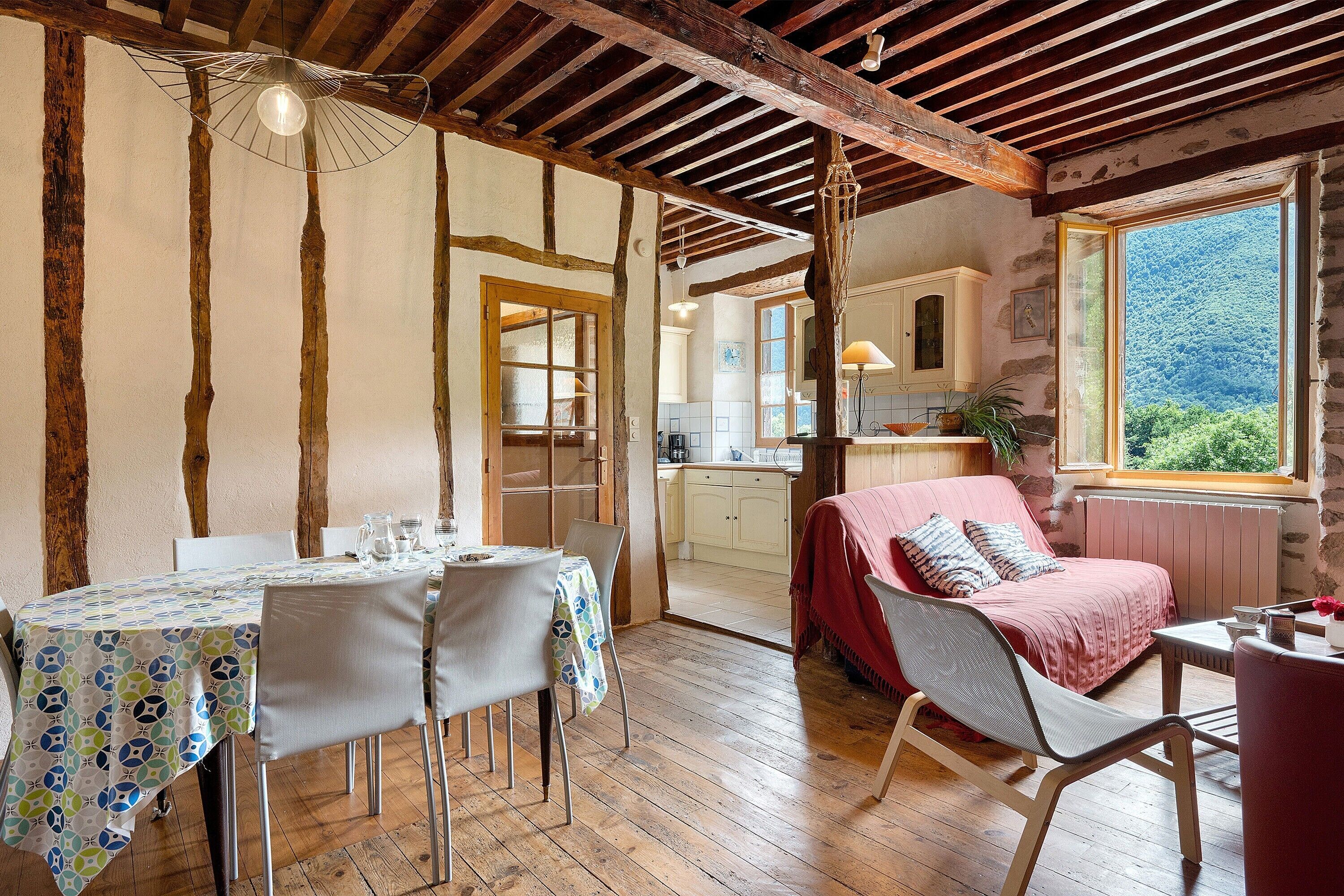 Une pièce rustique comprenant une table à manger, des chaises, un lit recouvert d’une couverture rouge, une fenêtre donnant sur un paysage verdoyant et, au fond, une cuisine.