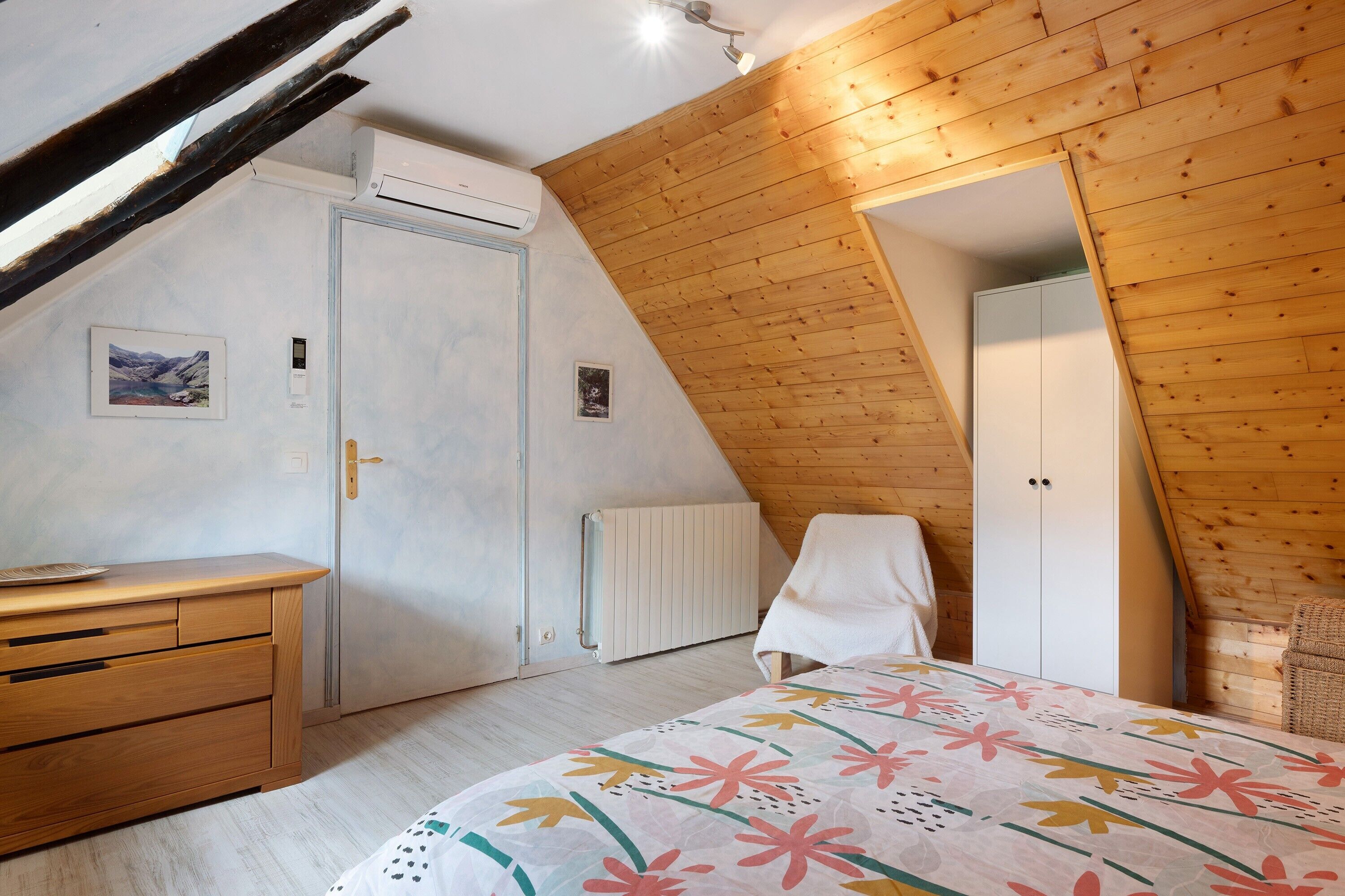 A bedroom with a sloped ceiling, wooden walls, a bed with a floral bedspread, a dresser, a radiator, and a door.