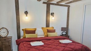 Ein Schlafzimmer mit roter gesteppter Bettdecke, Holzbalken an der Decke, zwei Wandleuchten, einem Nachttisch mit einer Uhr und einer Tür rechts.