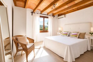 Ein Schlafzimmer mit einem großen Bett, einem Korbstuhl, einem Fenster mit Vorhängen und einer Holzbalkendecke.