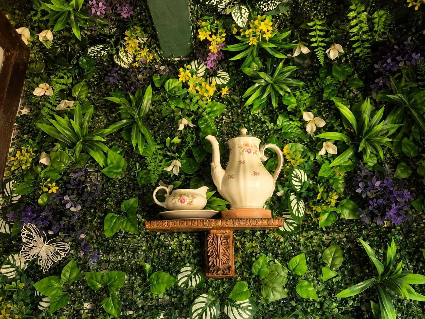 Un mur orné de différentes plantes et fleurs, une petite table avec une théière et une tasse, et un papillon décoratif.
