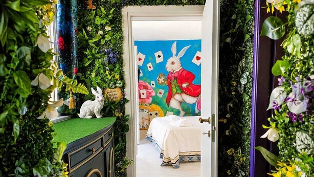 Ein Zimmer mit einem Bett, einem Schminktisch und einer Tür, die in einen anderen Raum mit einem Hasenbild führt.