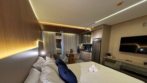 Quarto de hotel com cama, televisão, escrivaninha e cozinha compacta.