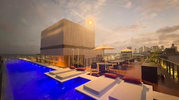 A rooftop pool area with lounge chairs and umbrellas, overlooking a city skyline.