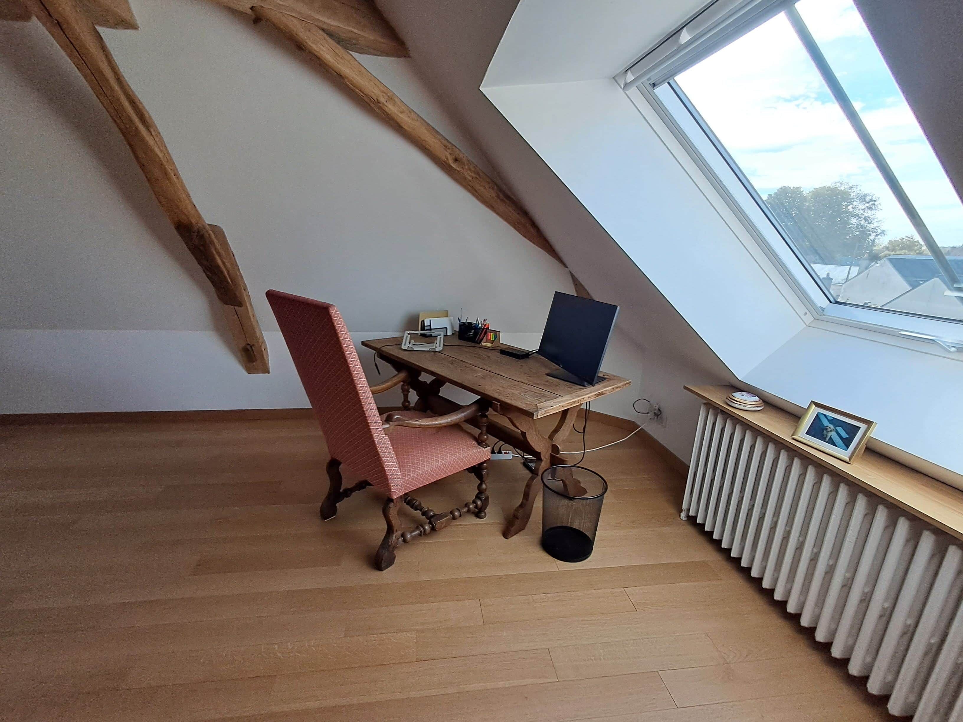 A room with a sloped ceiling, a wooden desk with a laptop, a red chair, and a radiator.