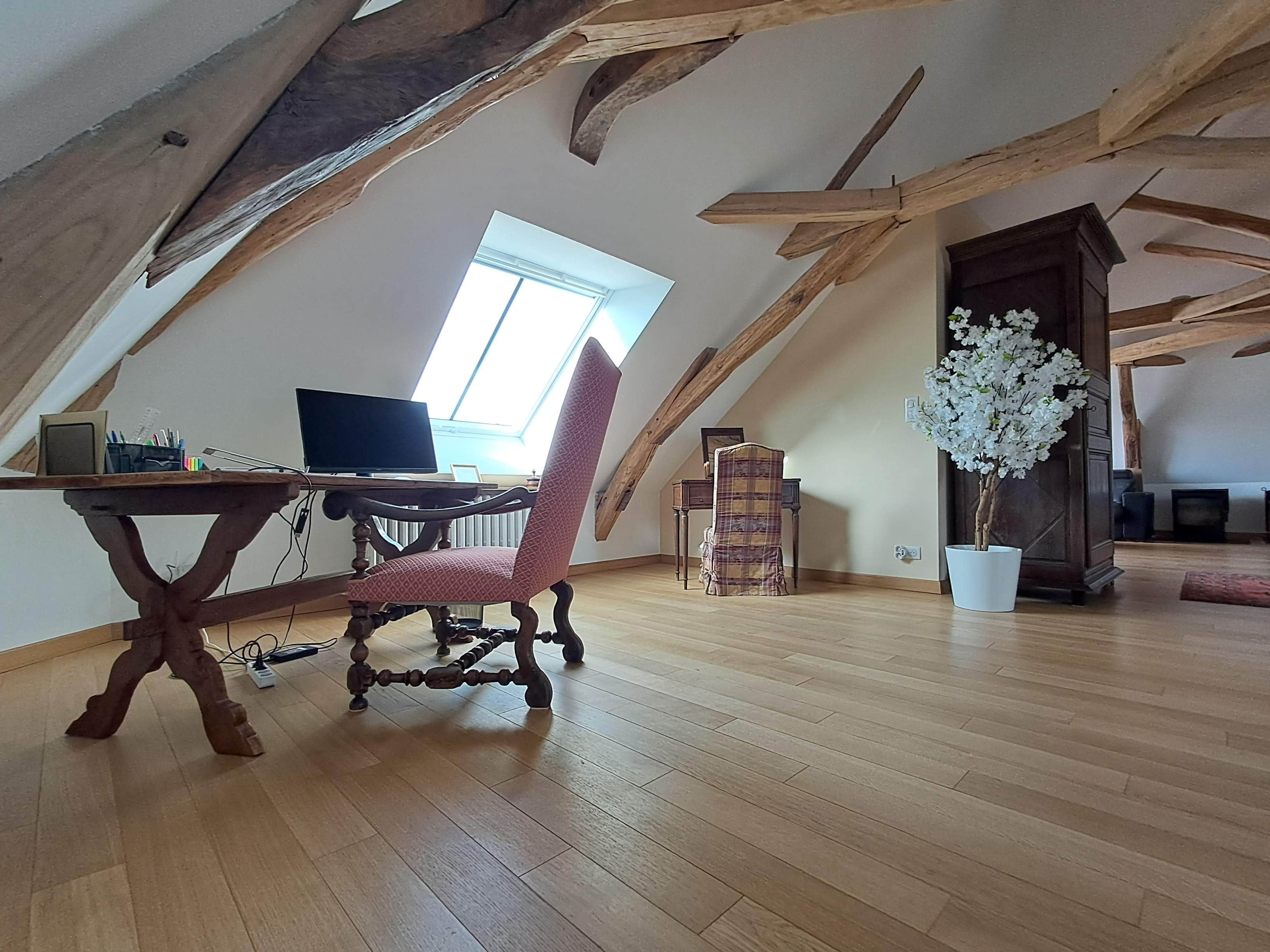 Un dormitorio en el ático con vigas de madera, un escritorio con una computadora, una silla roja y una planta en maceta.