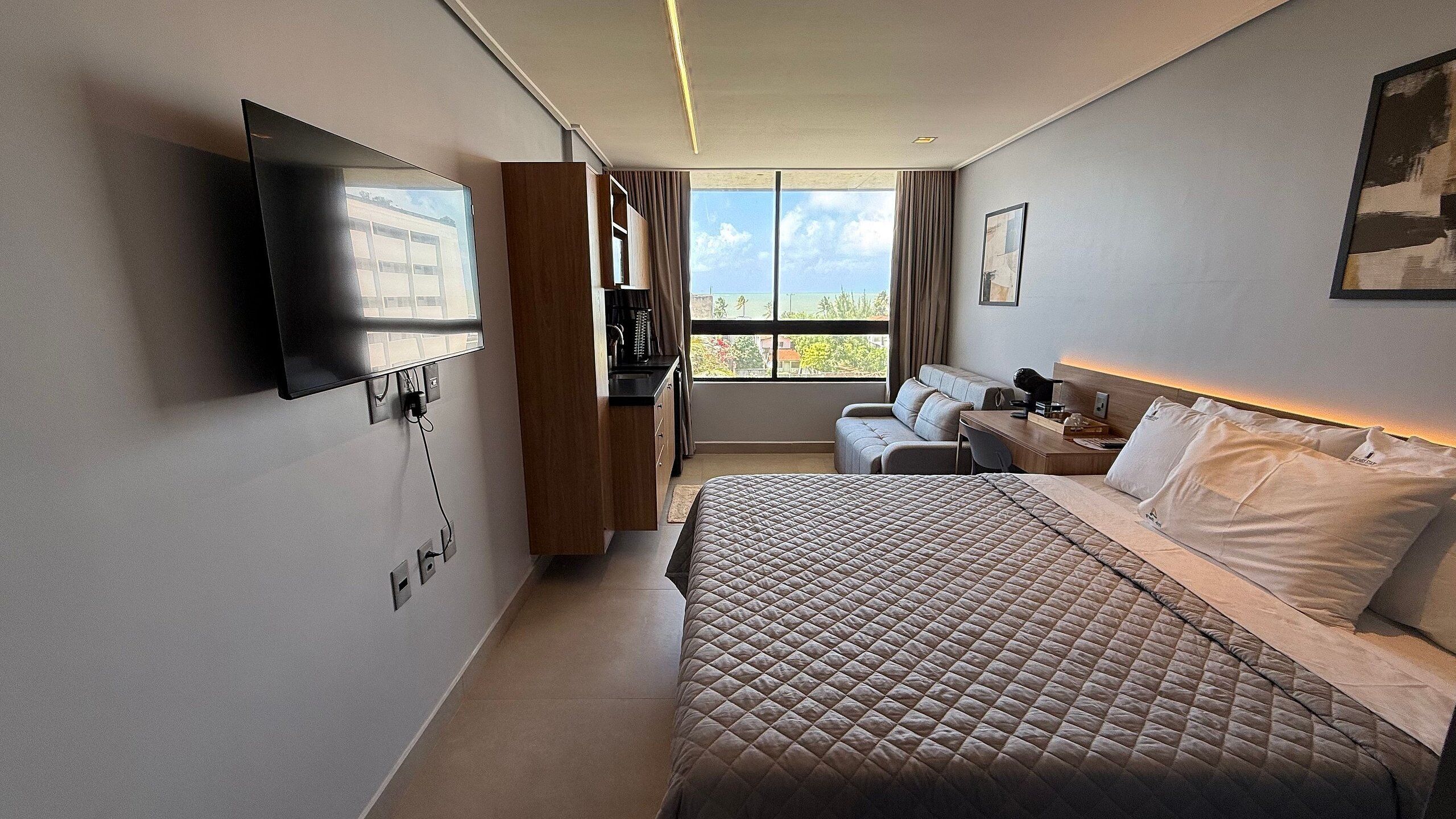 A hotel room with a bed, a TV mounted on the wall, a desk with a chair, a window with a view, and a sofa.