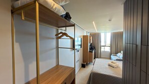 Ein Hotelzimmer mit Etagenbett, Schreibtisch, Kleiderschrank, Fernseher, einer kleinen Küchenzeile, einem Bett mit Handtüchern und einem Fenster mit Vorhängen.