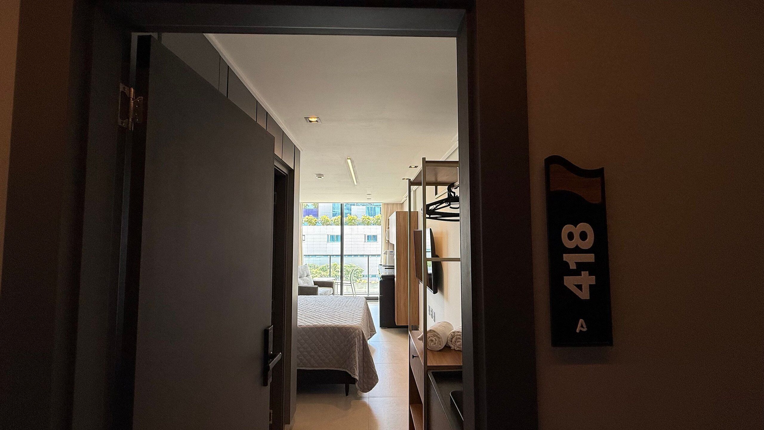 A hotel room with a bed, a desk, a chair, and a wardrobe. There is a window with a view of the outside.