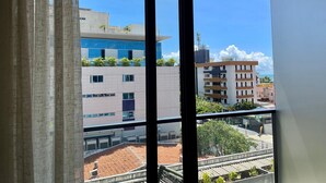 Blick durch ein Fenster mit vertikalen Jalousien. Man sieht ein mehrstöckiges Gebäude mit Balkonen, die mit Pflanzen bepflanzt sind, und einen strahlend blauen Himmel.