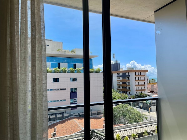Vista attraverso una finestra con persiane verticali, che mostra un edificio con diversi piani, balconi con piante e un cielo azzurro intenso.