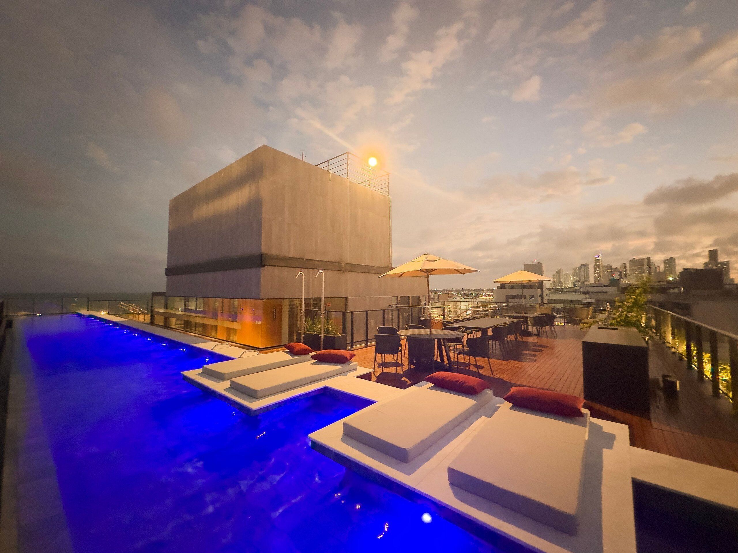 A rooftop pool area with lounge chairs and umbrellas, overlooking a city skyline.