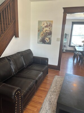 A living room with a dark leather sofa, a wooden staircase, a painting of flowers on the wall, and a dining area with a table and chairs.