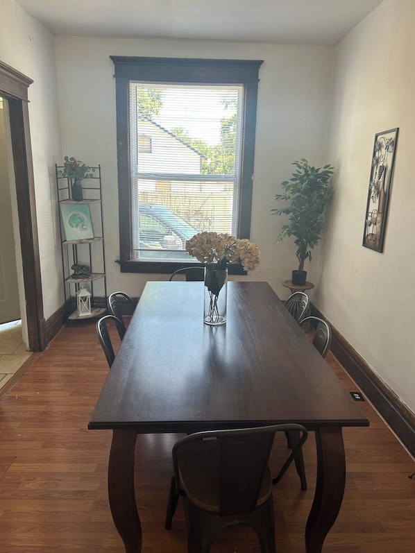 A dining room with a wooden table, chairs, a vase with flowers, a window with blinds, and a potted plant.