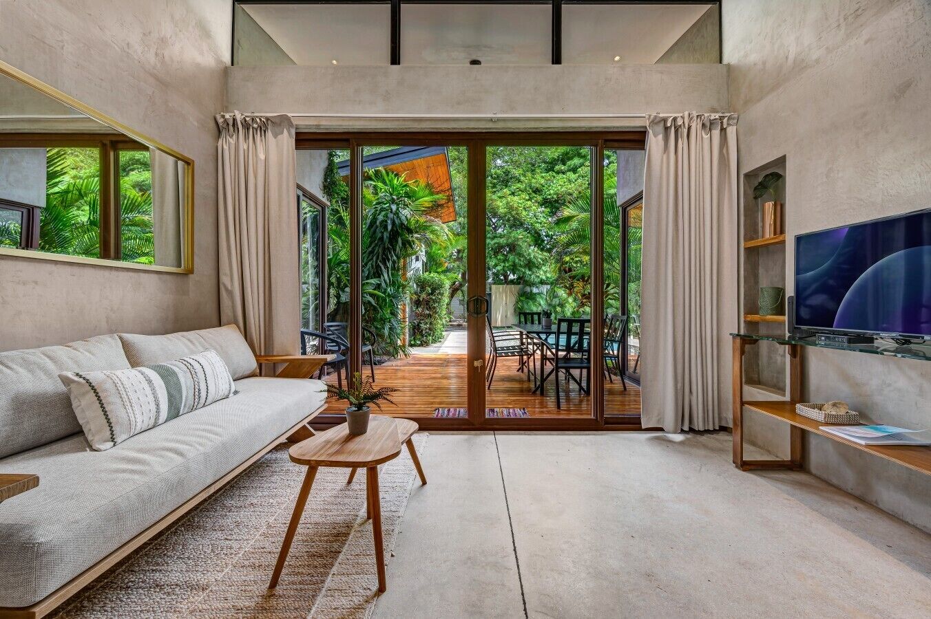 A modern living room with a sofa, a coffee table, a TV, and a sliding glass door leading to an outdoor patio with a pool and greenery.