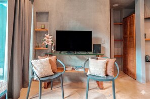 Ein Wohnzimmer mit einem Flachbildfernseher, zwei Sessel mit Kissen, einem Glastisch und einem Bücherregal.