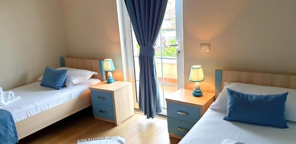 A hotel room with two single beds, bedside tables with lamps, a window with curtains, and a view of the outdoors.