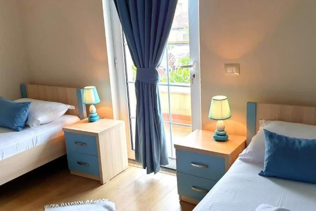 Habitación de hotel con dos camas individuales, mesitas de noche con lámparas, una ventana con cortinas y vistas al exterior.