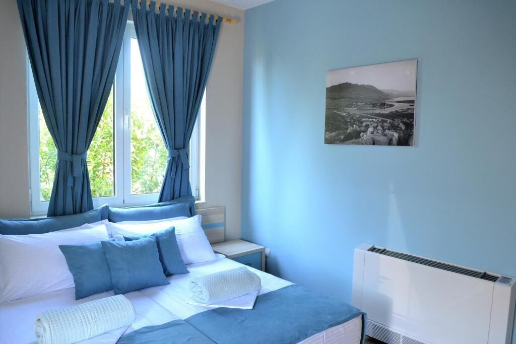 A bedroom with a bed, curtains, a nightstand, a wall-mounted picture, and an air conditioning unit.