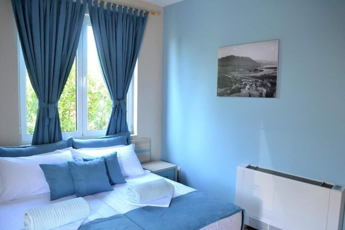 Un dormitorio con cama, cortinas, una mesita de noche, un cuadro en la pared y un equipo de aire acondicionado.