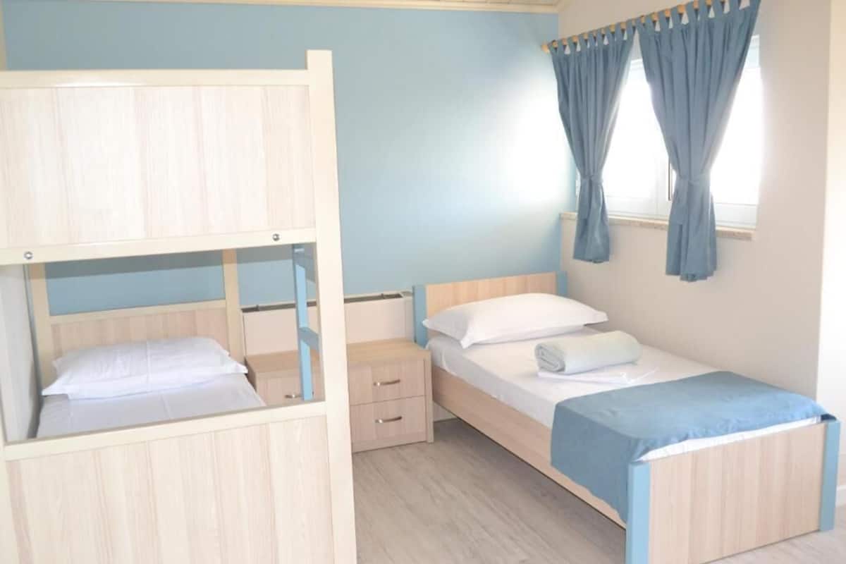 Una habitación con literas, una cama individual, una mesita de noche y una ventana con cortinas.