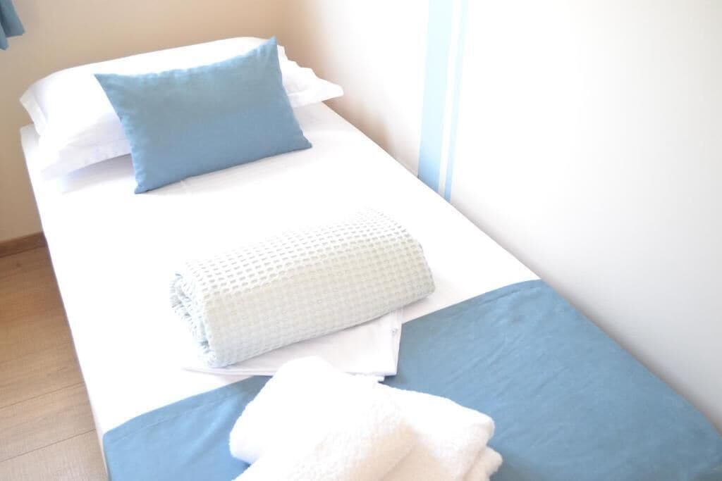 A single bed with a blue pillow, a white pillow, a white towel, and a rolled white towel.