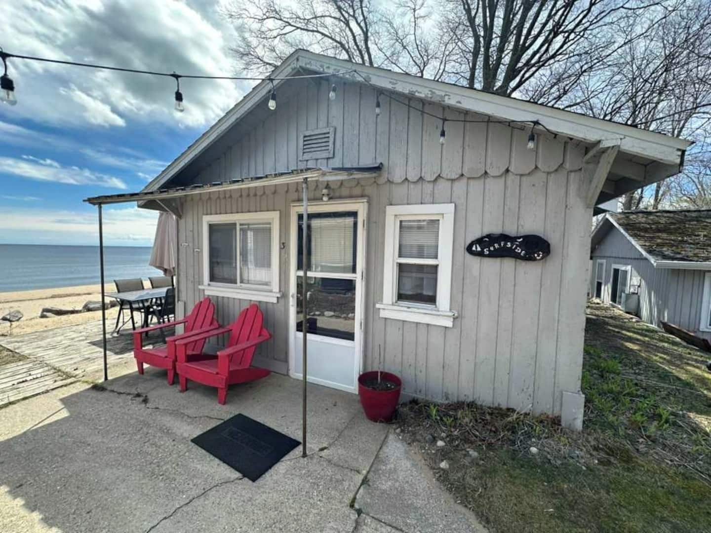 Uma cabana pequena com vista para a praia, um alpendre com cadeiras vermelhas e uma placa indicando 'Surfside'.