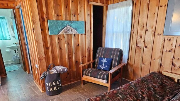 Una habitación con paneles de madera, una cama, un sillón con cojín, un bolso y un cuadro en la pared.