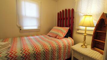 Un dormitorio con cama, mesita de noche, lámpara, ventana con persianas y estante en la pared.