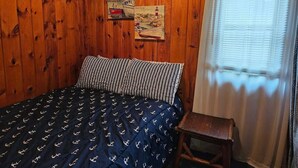 Una habitación con paredes de madera, una cama con colcha estampada, un taburete de madera y una ventana con persianas.