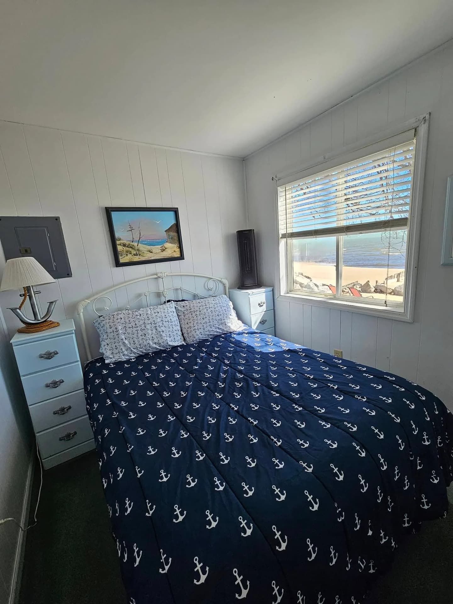 A bedroom with a bed, a nightstand, a lamp, a dresser, a window with a beach view, and a painting on the wall.