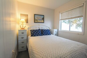 A bedroom with a bed, a nightstand with a lamp, a dresser, a painting on the wall, and a window with blinds.