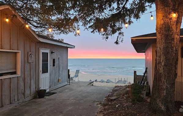 A wooden building with a door and windows, a paved path leading to a beach, and string lights above.