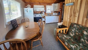 Interior de una cabaña de madera con una mesa de comedor, sillas, un sofá, una kitchenette y un ventilador de techo.