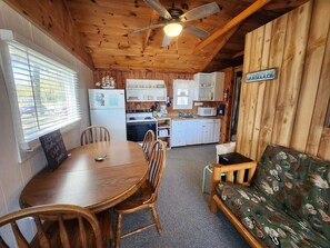 L’intérieur d’un chalet en bois comprend une table à manger, des chaises, un canapé, un coin cuisine et un ventilateur de plafond.