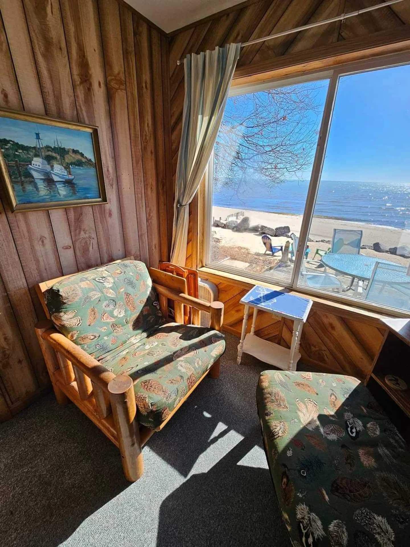 A room with wooden walls, a large window with a view of a beach, a wooden chair, a small table, and a patterned sofa.