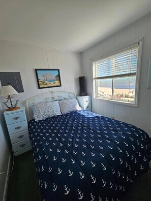 A bedroom with a bed, a nightstand, a lamp, a picture on the wall, and a window with a view of the beach.