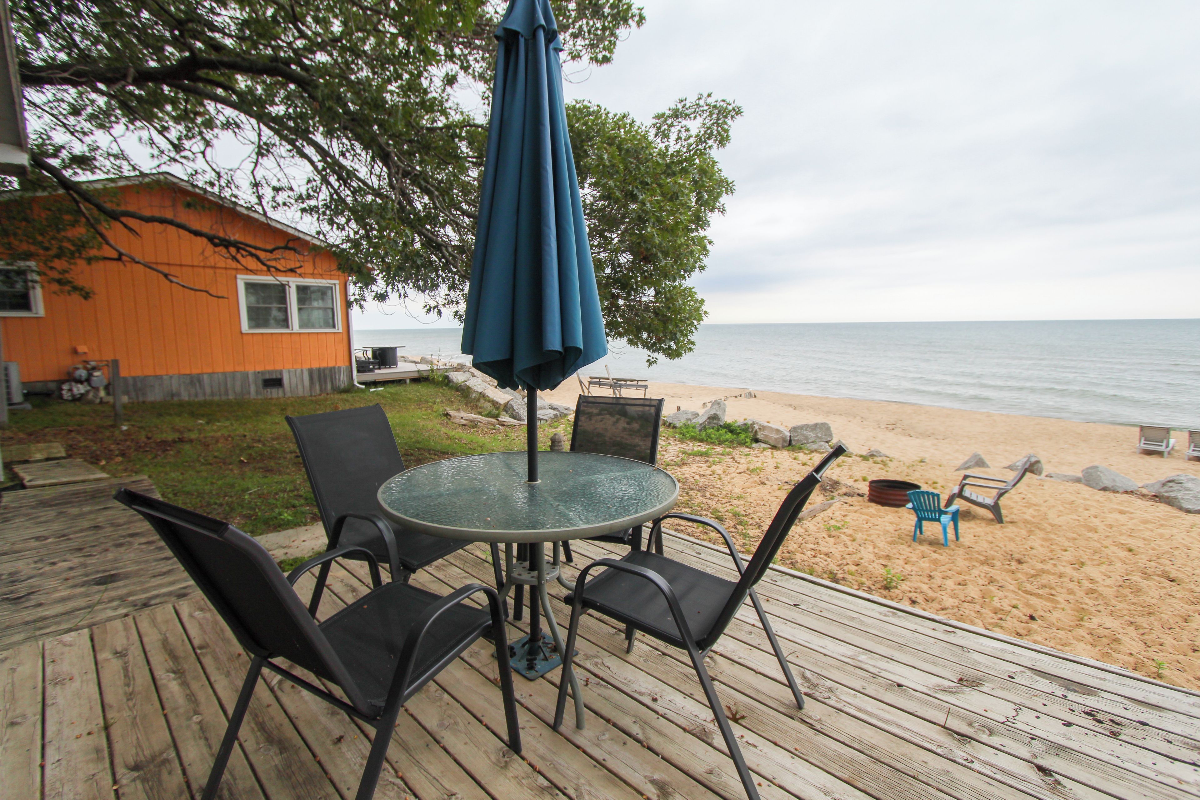 Une terrasse avec une table ronde et des chaises, donnant sur une plage de sable et une étendue d’eau.