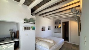 A bedroom with a bed, shelves with dishes, a desk, and a door leading to another room. - #HH11 Guestroom for two in Katavati (Sifnos)