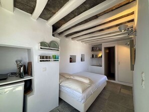 A bedroom with a bed, shelves with dishes, a desk, and a door leading to another room. - #HH11 Guestroom for two in Katavati (Sifnos)