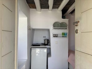 A compact kitchenette with a refrigerator, a kettle, and shelves with dishes and cups. - #HH11 Guestroom for two in Katavati (Sifnos)