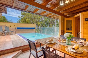 A poolside dining area with a table set for two, a wooden ceiling, and a view of the pool.