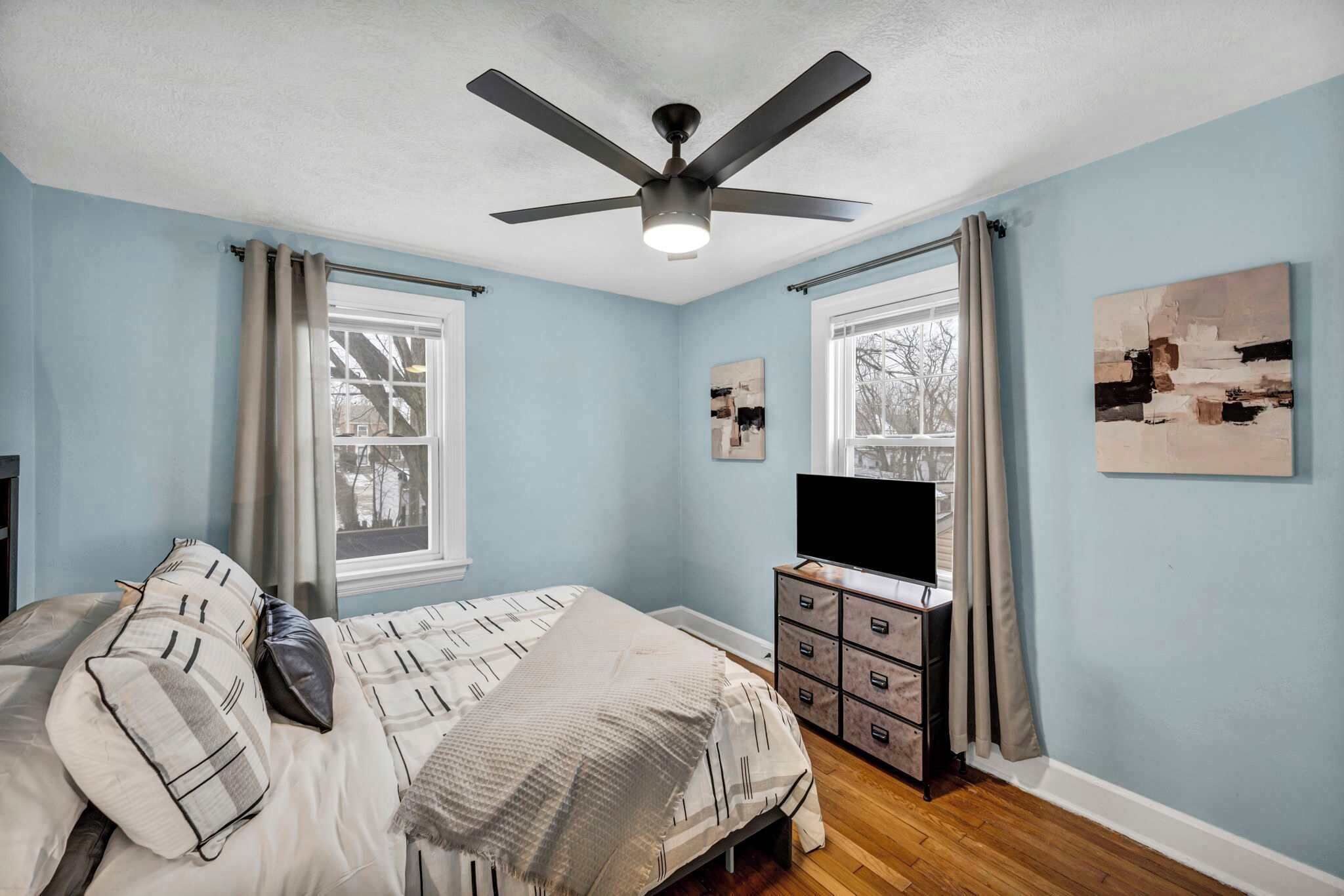 A bedroom with a bed, a ceiling fan, a TV, and artwork on the walls.